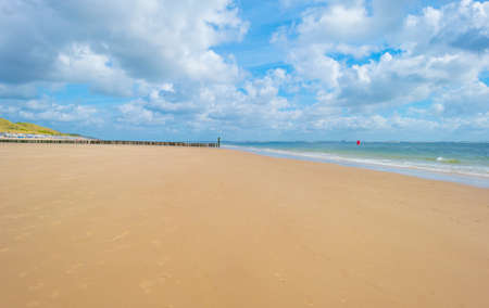 Sunlit waves and a wooden breakwater on the yellow sand of a sunny beach along a sea under a blue...