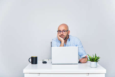 Business man teleworking from home with a laptop sending a message with his cell phone. Bald man with glassesの写真素材