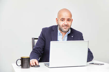 Business man teleworking from home with a laptop sending a message with his cell phone. Bald man with glassesの写真素材