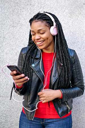 A portrait of a smiling African-American woman with a cell phone is chatting or listening to music on the streets of the city. The hair is braided into dreadlocks.の写真素材