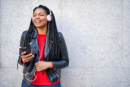 A portrait of a smiling African-American woman with a cell phone is chatting or listening to music on the streets of the city. The hair is braided into dreadlocks.の写真素材