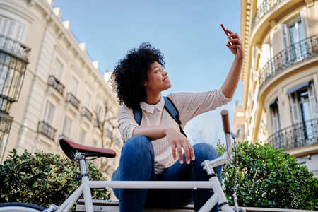 African American woman taking a self-portrait with her smart phone. Sitting on a bench with her...
