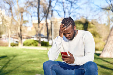 African American man sitting on a park bench looking at his cell phone. Concerned young man. Concept...