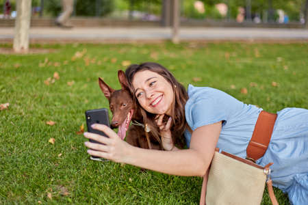Pretty young woman taking a selfie with her pharaoh dog lying on the grass in a park.の写真素材