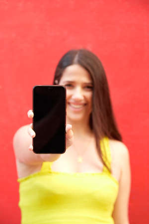 Young girl smiling showing mobile phone with yellow top on red background. Selective focus on smart...