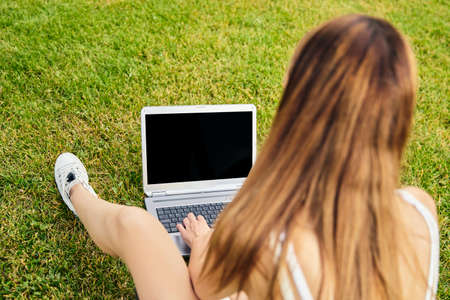 Rear view of young woman , sitting on the grass, chatting with her family using a laptop, studying, shopping online, surfing the net. Concept of people and technology.の写真素材