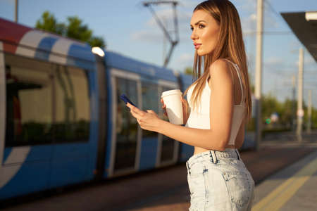 Young woman waiting for city train and talking on phone with coffee in hand. Passenger at streetcar stop with smartphone. Concept of public transport and communication. High quality photo.の写真素材