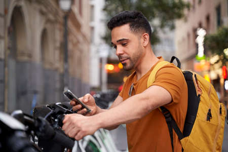 Positive young Latino man stands by the parking lot with a rental bike and uses a smartphone with a smile on his face. Eco-friendly transport conceptの写真素材