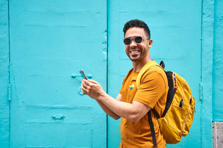 Cheerful young Latino looking at the camera with his backpack typing on his cell phone walking down the street, wearing orange t-shirt with lgtbi sticker and sunglasses.の写真素材