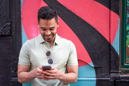 Cheerful young latin guy texting, checking social networks, chatting on his smart phone on colorful...