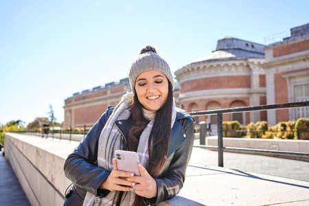 Confident young female millennial with long dark hair in warm knitted hat and leather jacket smiling...