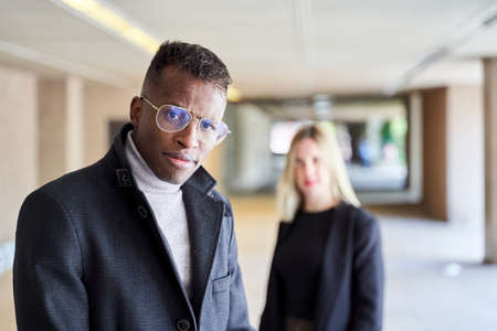 African American man in stylish coat and glasses looking at camera while standing in modern passage...