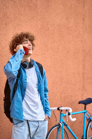 Smiling curly-haired young woman standing on wall outdoors talking on cell phone next to her blue bicycle.の写真素材