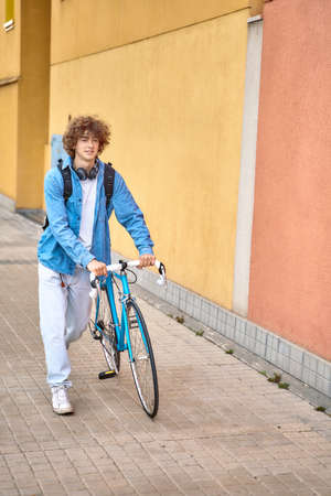 Young curly-haired student with headphones walking to college holding bicycle with hands.
