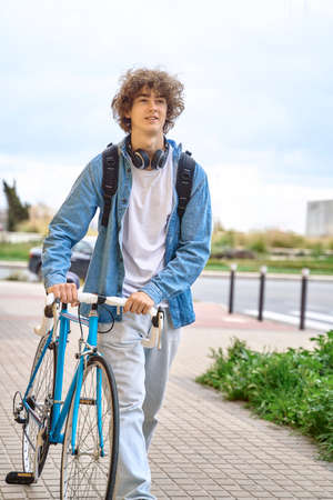 Young curly-haired student with headphones walking to college holding bicycle with hands.の写真素材