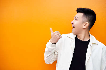 Happy Asian male in white jacket and black t shirt smiling for camera and showing thumb up gesture...
