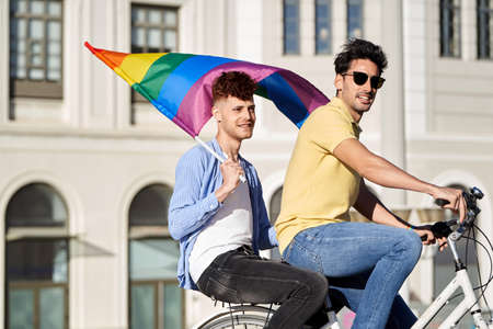 Young gay couple holding a gay pride flag while riding a bike outdoors. LGBT, relationship and equal...