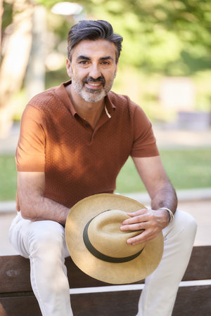 Relaxed middle-aged man holding a hat and looking at camera while sitting on a bench in a park.の写真素材