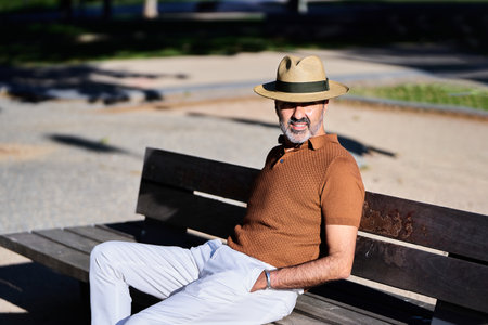 Man with a hat looking away while relaxing sitting on a bench in the park.の写真素材