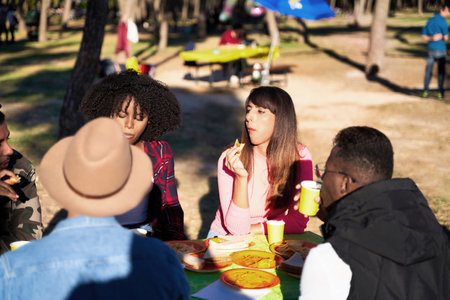 Group of multi-ethnic friends looking at camera and smiling while enjoying having a picnic together sitting on a table outdoors in a park. Friendship concept.の写真素材