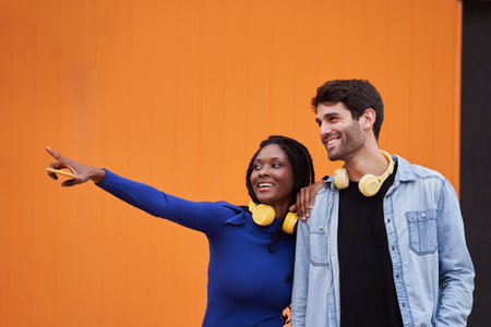 Man and woman with wireless headphones pointing away and smiling while standing outdoors.