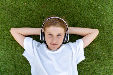 boy with headphones listening to music lying on the grass in the park looking at the camera