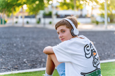 Teenage boy looking at the camera while listening to music with headphones outdoors in a park....