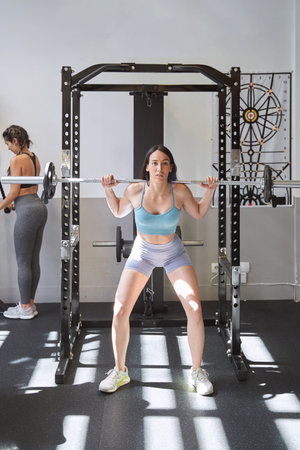 A powerful and determined young woman performing a shoulder press with a heavy barbell during her intense workout session.の写真素材