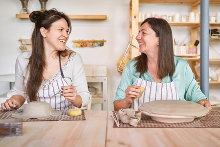 Two women in aprons smiling while enjoying together in a ceramic workshop. Hobby and craft concept.