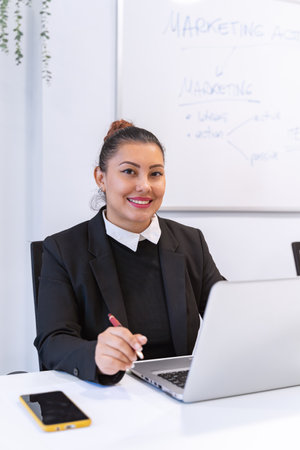 A cheerful Latina woman, working in an office, smiles and looks directly at the camera, radiating...