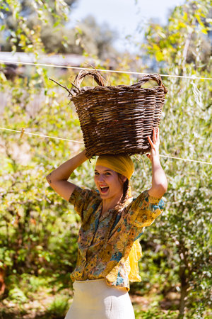 Farmer woman looking at the camera while carrying a wicker basket on her head outdoors in the garden.の写真素材