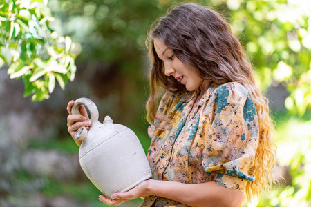 Young woman smiling while holding a clay jug standing outdoors in a garden.の写真素材