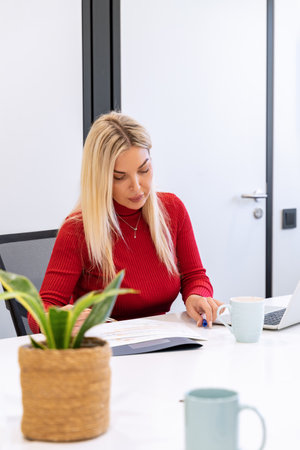 A focused businesswoman in a red sweater reviews charts and reports at her white office deskの写真素材