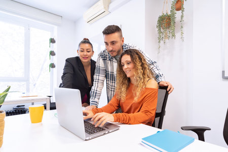A multi-ethnic team of three collaborates around a laptop in a bright office adorned with green...