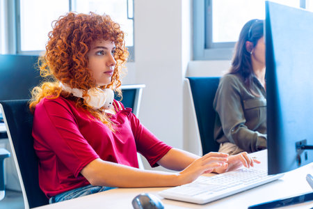 Red-haired woman with headphones working on computer in a bright office setting.