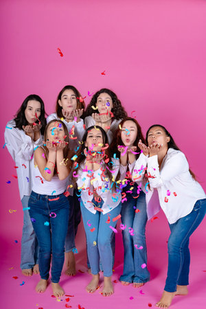 Joyful group of young women in white shirts and jeans blowing colorful confetti on a pink background.の写真素材