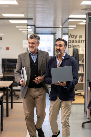 Two mature executives walking together in the office, one with laptop and one with documents.の写真素材