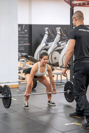 Concentrated female athlete performs a barbell pull with meticulous guidance from her fitness coach in a gym setting.の写真素材