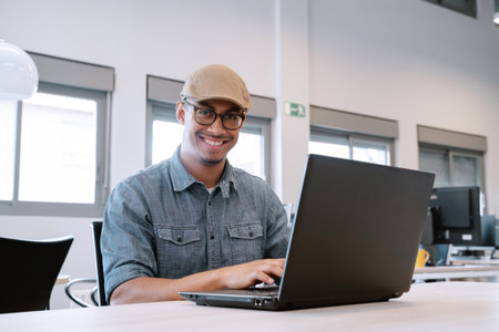 Cheerful Latino man with glasses and flat cap working on a laptop in a bright and spacious office, smiling at the camera.の写真素材