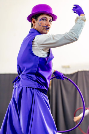Clown with purple makeup and costume performing with hula hoop, entertaining audience under circus tentの写真素材