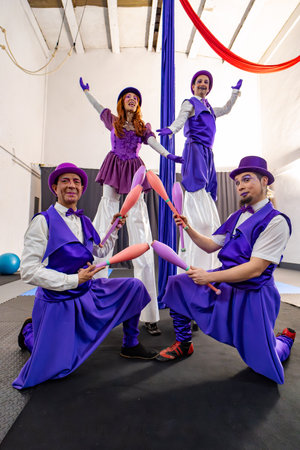 Four circus performers in vibrant purple costumes showcase their juggling and stilt walking skills inside a training gymの写真素材