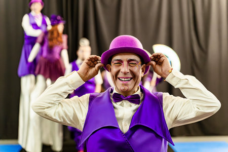 Cheerful circus performer in vibrant purple costume adjusts his hat, getting ready for a show, while other artists wait in the backgroundの写真素材