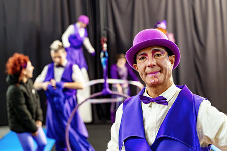 Smiling circus performer wearing purple costume and bowler hat posing backstage with colleagues practicing in the backgroundの写真素材
