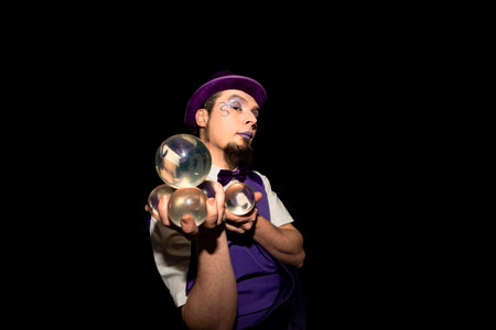 Juggler wearing purple makeup and holding four transparent juggling balls, preparing his performance in a dark environmentの写真素材