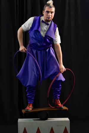 Male artist wearing purple costume balancing on seesaw while holding hoops, performing in circus showの写真素材