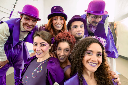 Group of smiling circus performers wearing purple costumes and makeup are posing together for a backstage photoの写真素材