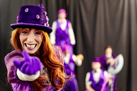 Redhead woman wearing purple costume and top hat, smiles and points her finger, with circus performers standing out of focus in the backgroundの写真素材