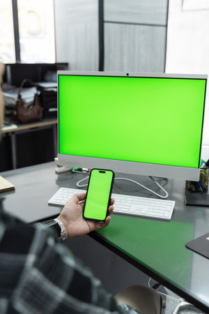 Office worker holding a smartphone with a vibrant green screen, positioned near a computer featuring a matching green screen on the deskの写真素材