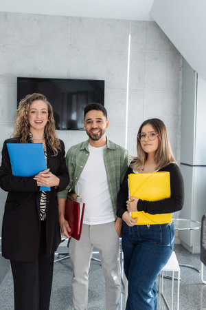 Three professionals, holding colorful folders, smiling in a modern office environment, suggesting teamwork and collaborationの写真素材