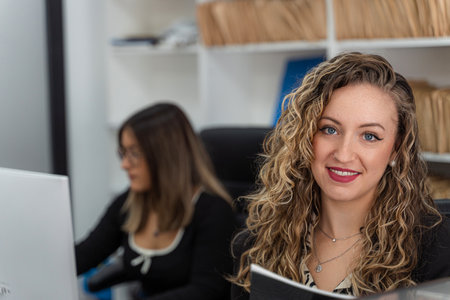 Portrait of a smiling businesswoman holding a folder in a modern office, with a colleague working on a computer in the backgroundの写真素材